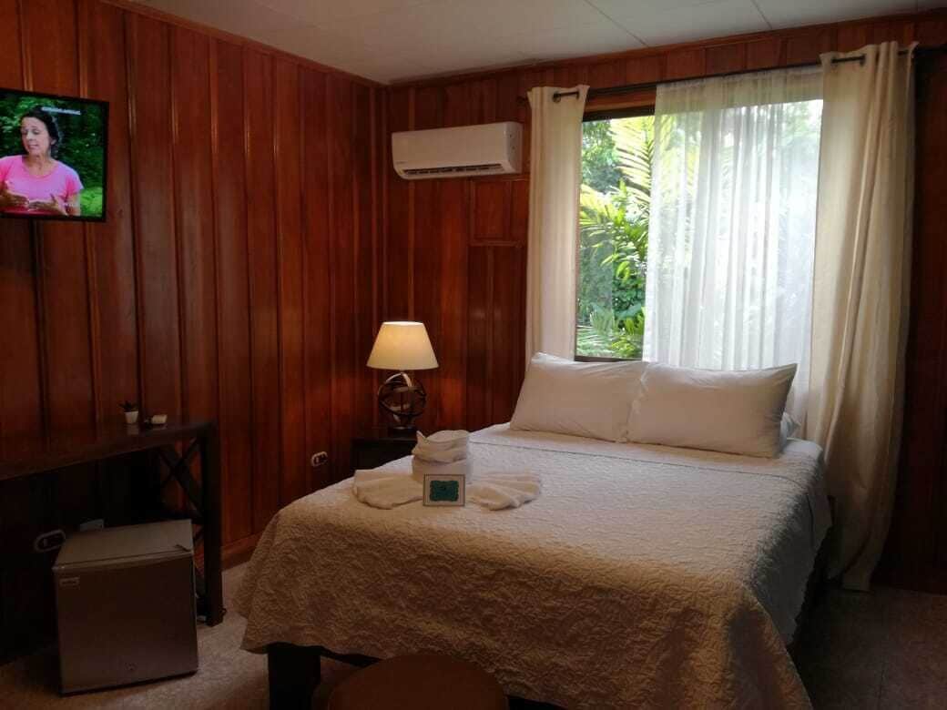 A hotel room with a wooden paneled wall, a bed with white linens, a nightstand with a lamp, a TV on the wall, and a window with curtains.