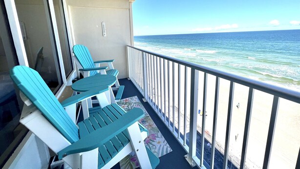 A balcony with two turquoise Adirondack chairs and a small table, overlooking a beach with waves.
