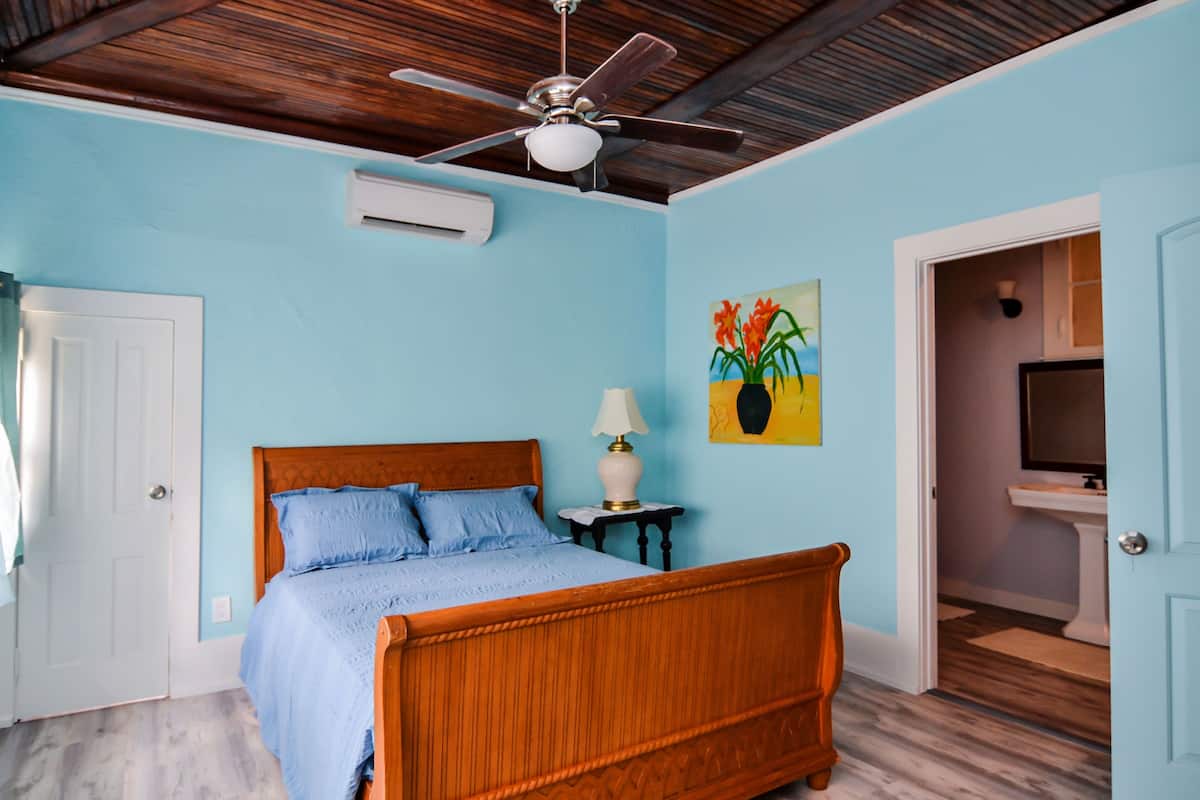 Quarto com cama de madeira, ventilador de teto, quadro na parede e banheiro visível através de uma porta aberta.