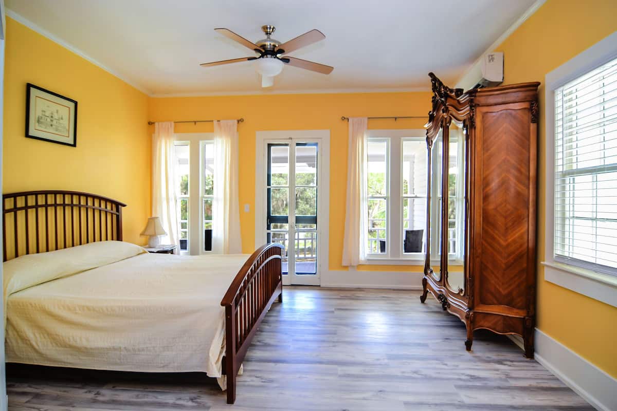 Quarto com cama, ventilador de teto, guarda-roupa de madeira e uma grande janela com persianas.