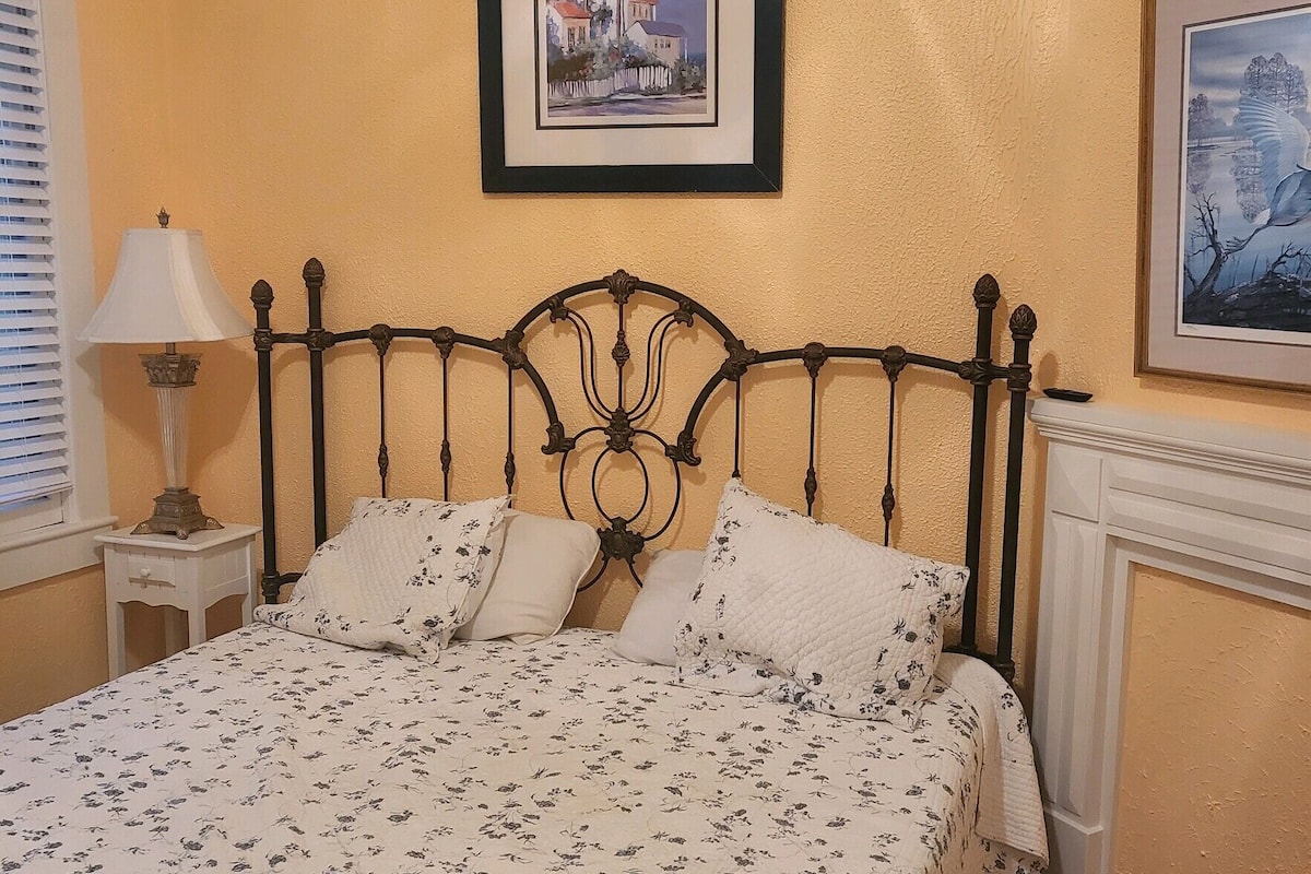 Uma cama bem arrumada com colcha florida, um quadro emoldurado acima e um abajur em uma mesa de cabeceira.