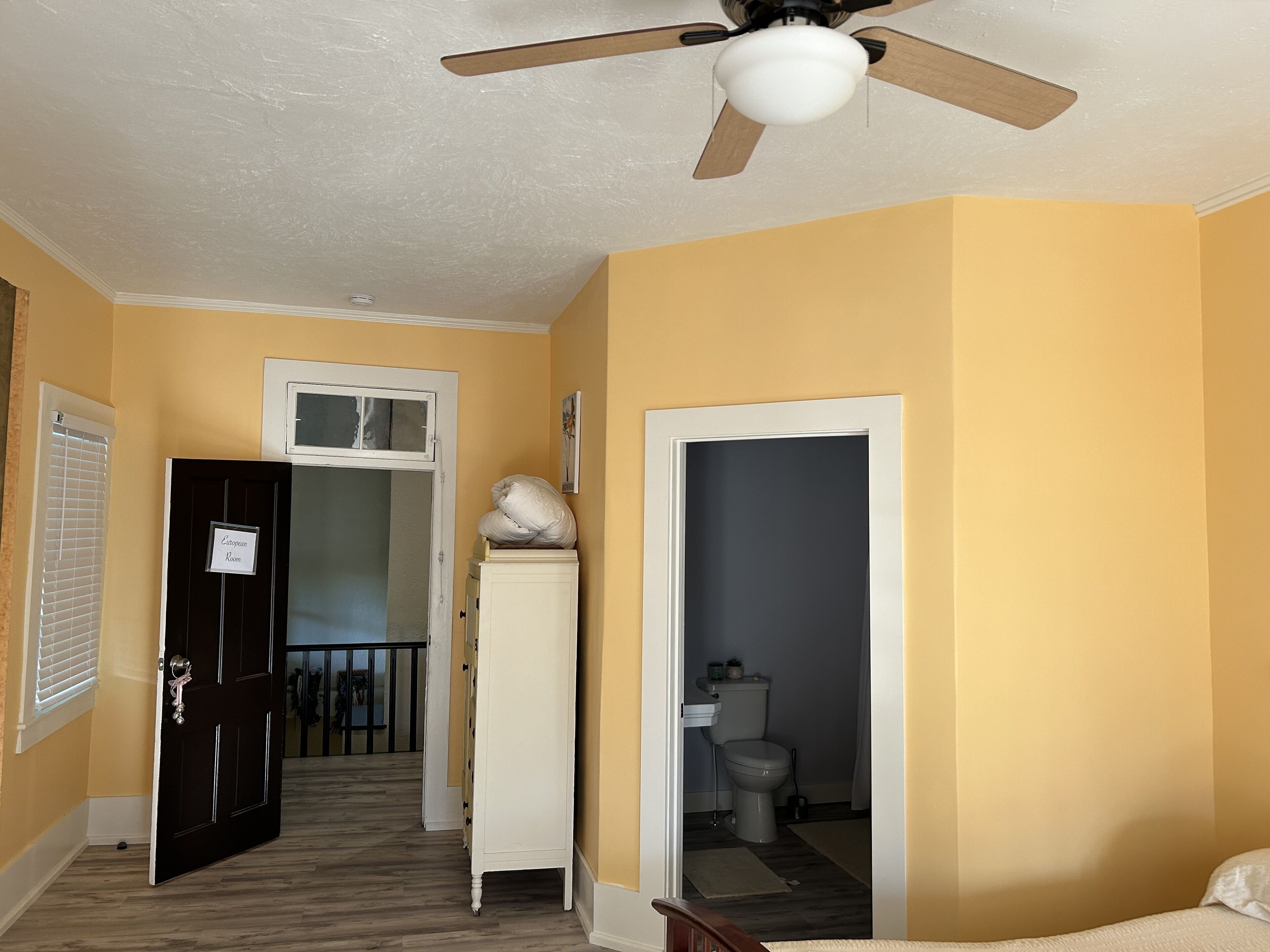 Une chambre d’hôtel avec un plancher en bois, un ventilateur de plafond, un lit, une salle de bain avec des toilettes et un lavabo, et une porte portant l’inscription « Chambre européenne ».