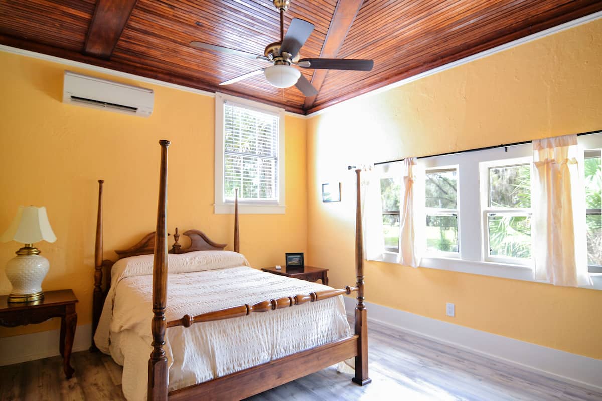Uma cama com dossel, cabeceira de madeira, ventilador de teto e janela com cortinas.