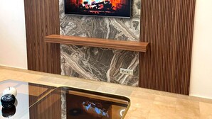 Een moderne woonkamer met een flatscreen televisie aan een marmeren muur, houten lambrisering en een glazen salontafel.