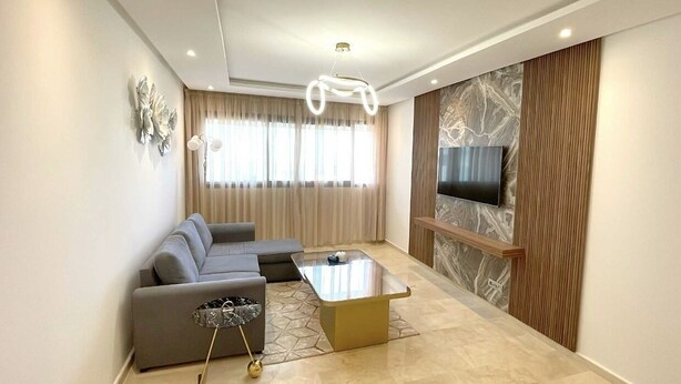 Een moderne woonkamer met een modulaire bank, een glazen salontafel, een flatscreen televisie die aan een marmeren muur is bevestigd, en een bijzondere hanglamp.