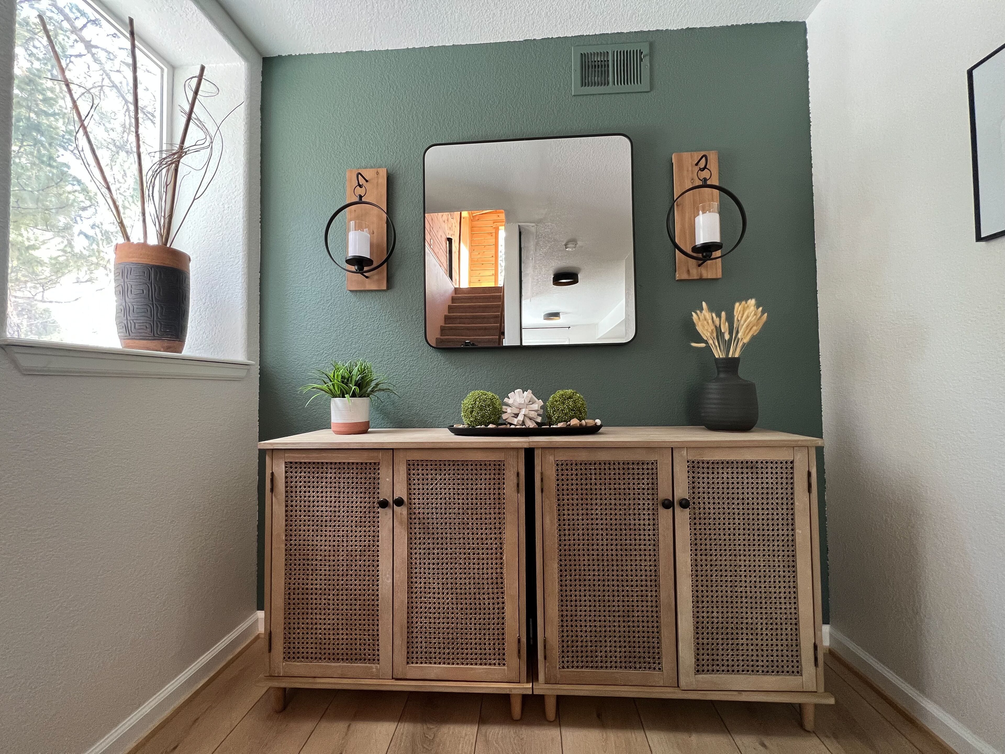 Un meuble en bois surmonté d’un miroir, avec deux appliques murales de chaque côté, et un pot de fleurs placé à gauche.