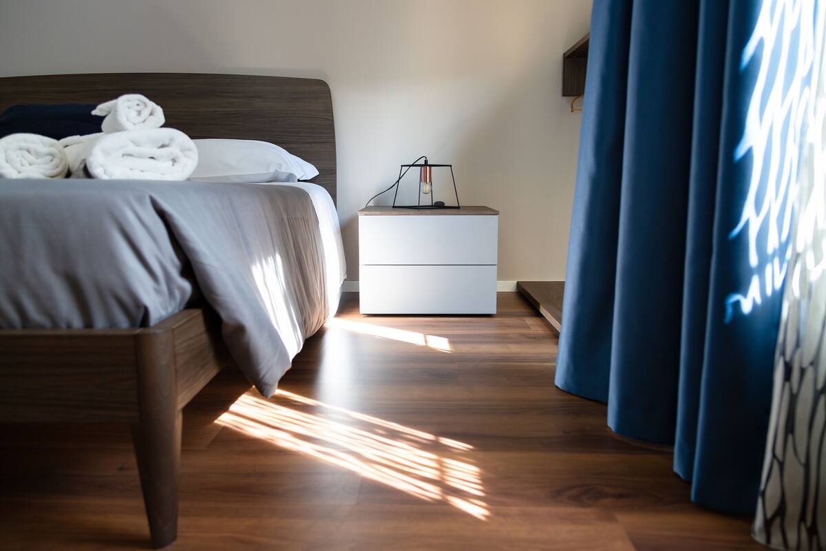 Una cama bien hecha con toallas enrolladas, una mesita de noche con una lámpara y una cortina azul.