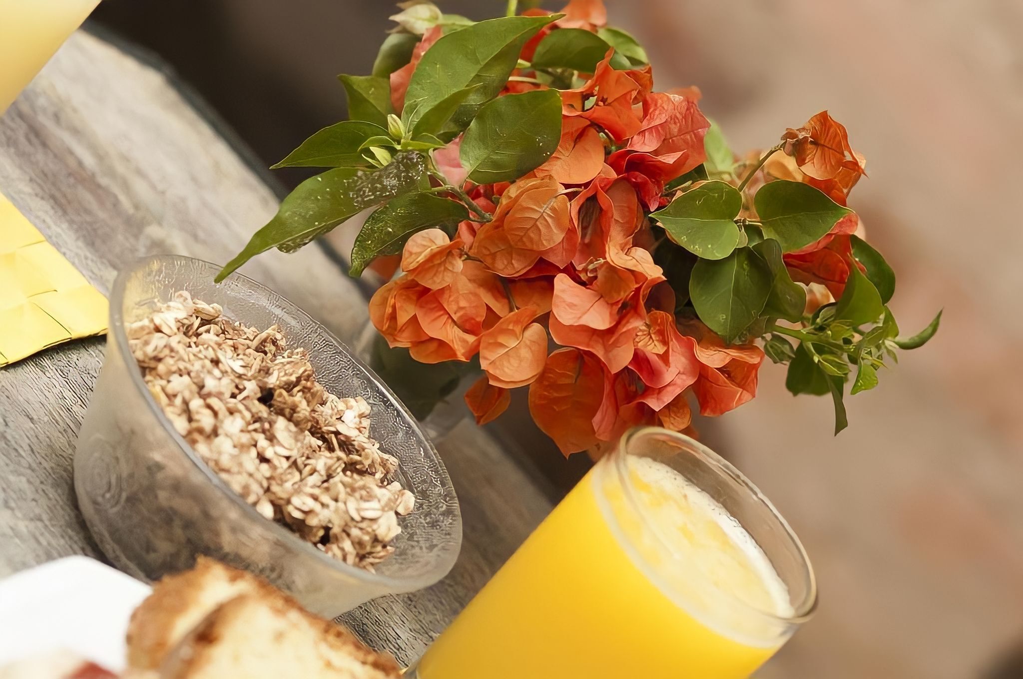 A breakfast setting with a bowl of granola, a glass of orange juice, and a bouquet of flowers.