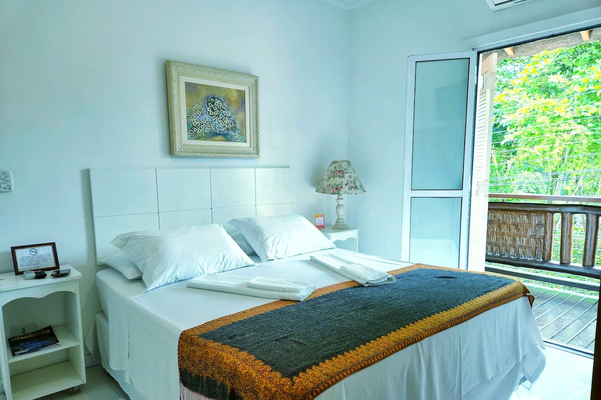 A hotel room with a bed, a nightstand, a lamp, a framed picture on the wall, and a balcony with a view of greenery.