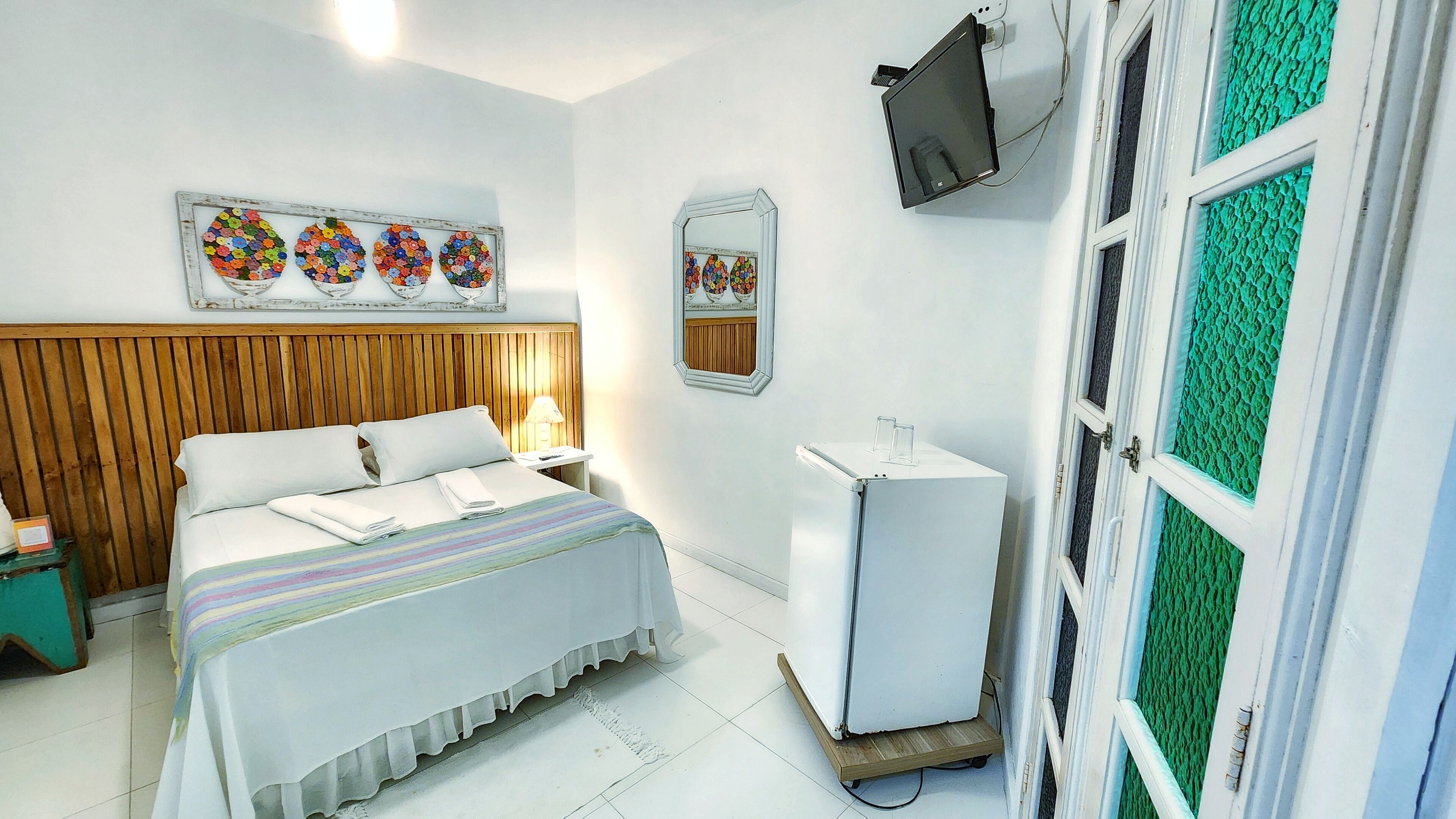 Camera d'albergo con una testiera in legno, un letto con lenzuola bianche, una piccola scrivania bianca, uno specchio e un televisore montato a parete.