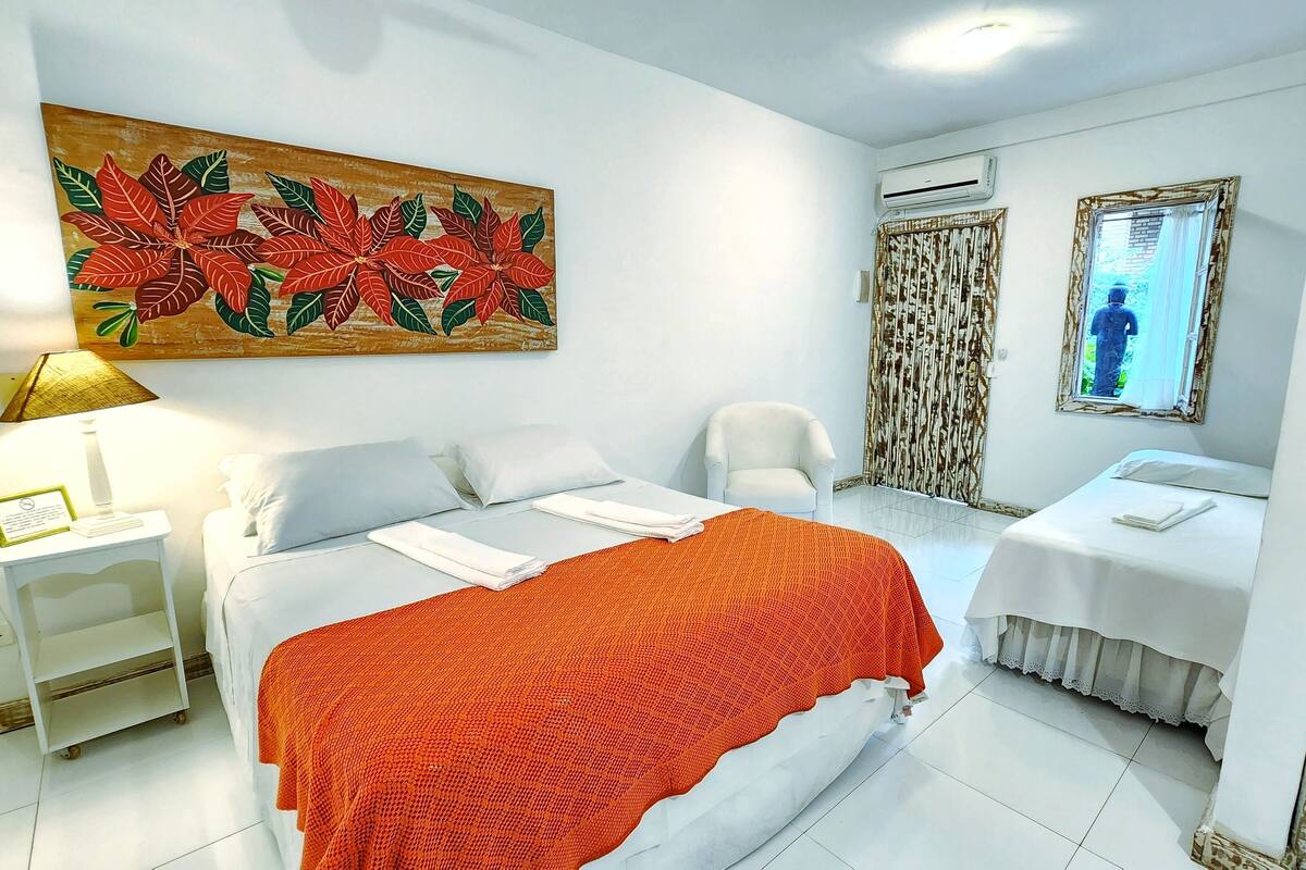 A hotel room with a large bed, a smaller bed, a white armchair, a side table with a lamp, and a painting of red flowers.