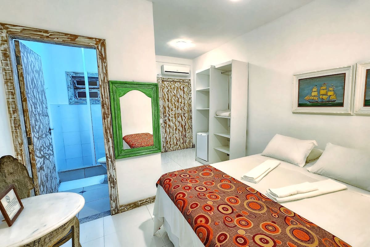 A hotel room with a bed, a wardrobe, a mirror, a small table, and a painting on the wall.
