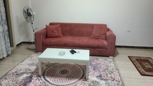 Una sala de estar con un sofá rojo, una mesa de centro de vidrio, un tapete estampado, un ventilador de pie y un cuadro en la pared.