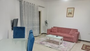 Una sala de estar con un sofá azul, un sofá rosa, una mesa de centro de vidrio, una mesa blanca, un tapete estampado y un ventilador.