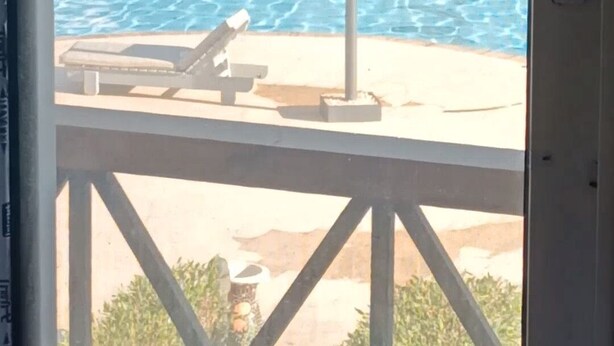Vista de una piscina con sillones de descanso y sombrillas, un edificio al fondo y un marco de puerta en primer plano.