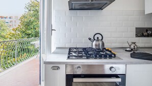 Cuina moderna amb fornell a gas, bullidor i forn, frontissa de rajola blanca i balcó amb barana metàl·lica.