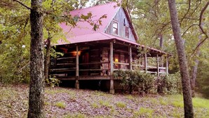 A rustic cabin with a red roof, surrounded by trees and a grassy area.