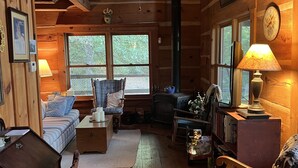 A cozy log cabin interior with a fireplace, wooden furniture, and a large window providing a view of the outdoors.