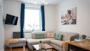 Uma sala moderna com um sofá seccional, uma mesa de centro, uma TV de tela plana na parede e um espaço de jantar com uma mesa posta para dois.