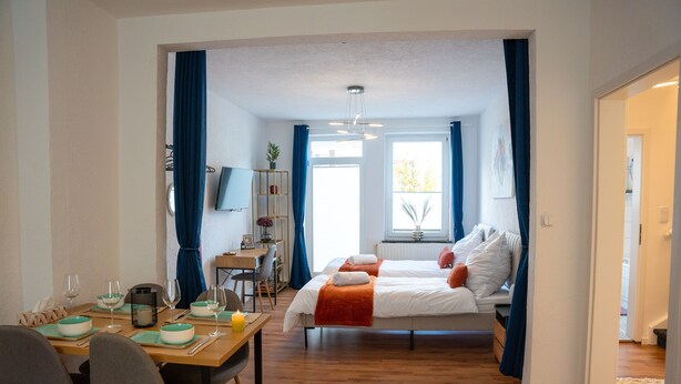 Quarto moderno com cama, mesa de cabeceira e escrivaninha com cadeira. Há uma janela com cortinas, uma planta e um quadro na parede.