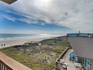 Une vue sur la plage depuis une terrasse en bois dotée d’une balustrade, donnant sur l’océan et une côte lointaine parsemée de bâtiments.