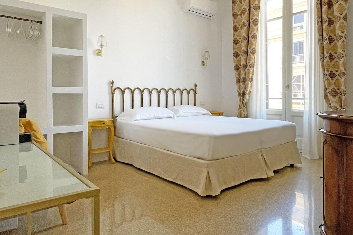 A bedroom with a bed, a desk, and a chair. There is a wardrobe with shelves, an air conditioning unit, and a window with curtains.