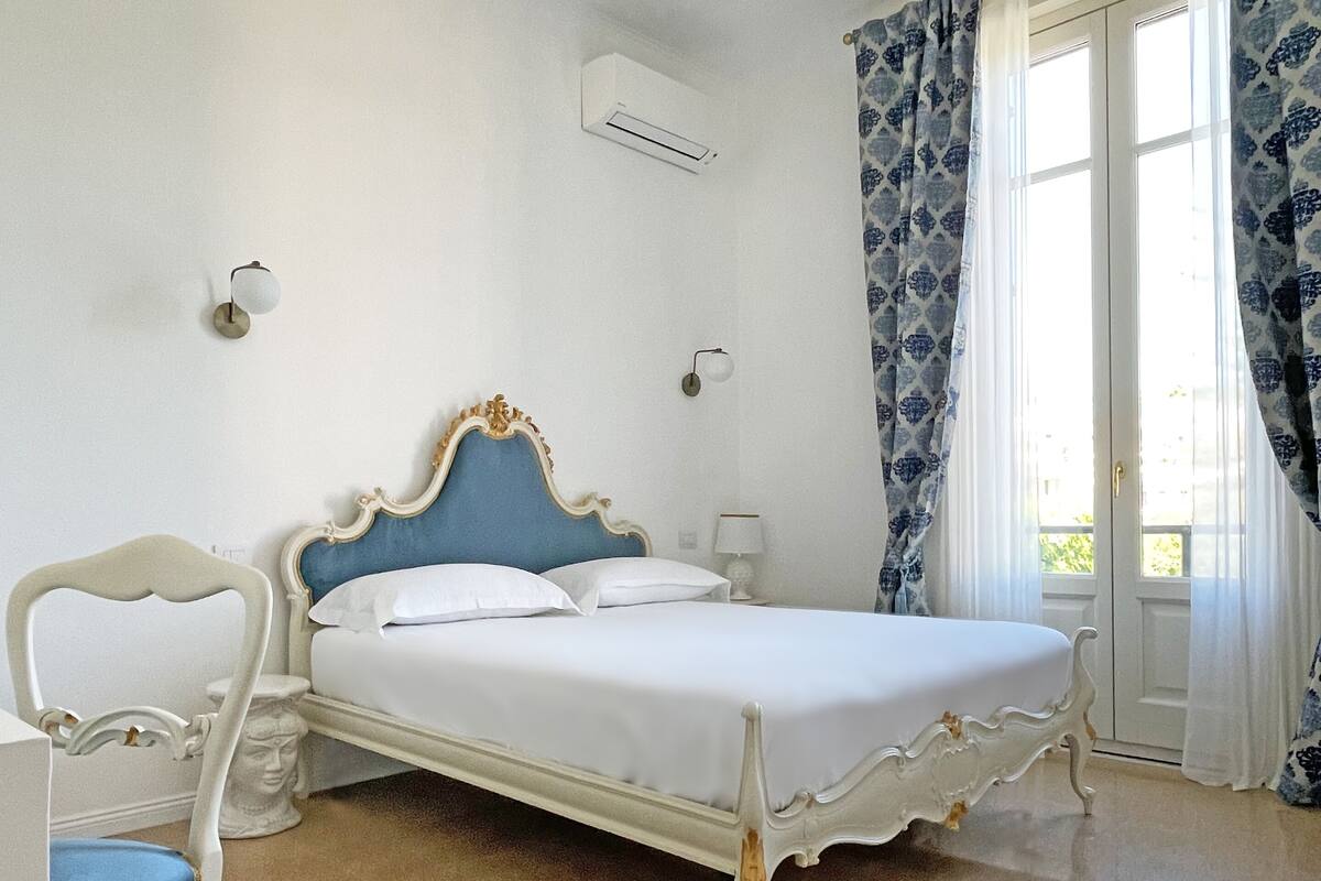 A bedroom with a white bed, a chair, a toilet, and a window with curtains.