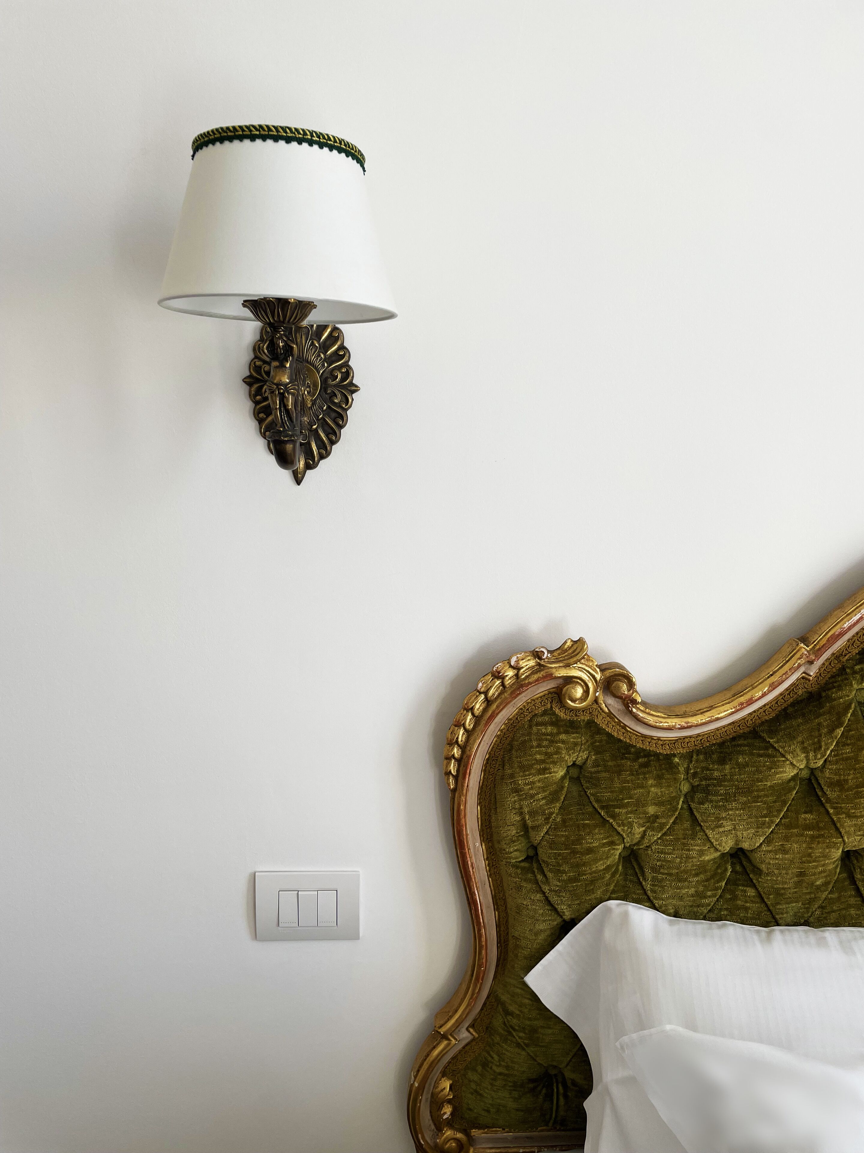 Una cama con cabecera tapizada de verde, una lámpara de pared con base decorativa y un interruptor de luz en la pared.
