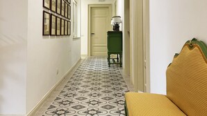 Un couloir avec des carreaux de sol à motifs, une petite table d’appoint verte avec une lampe et un banc jaune.