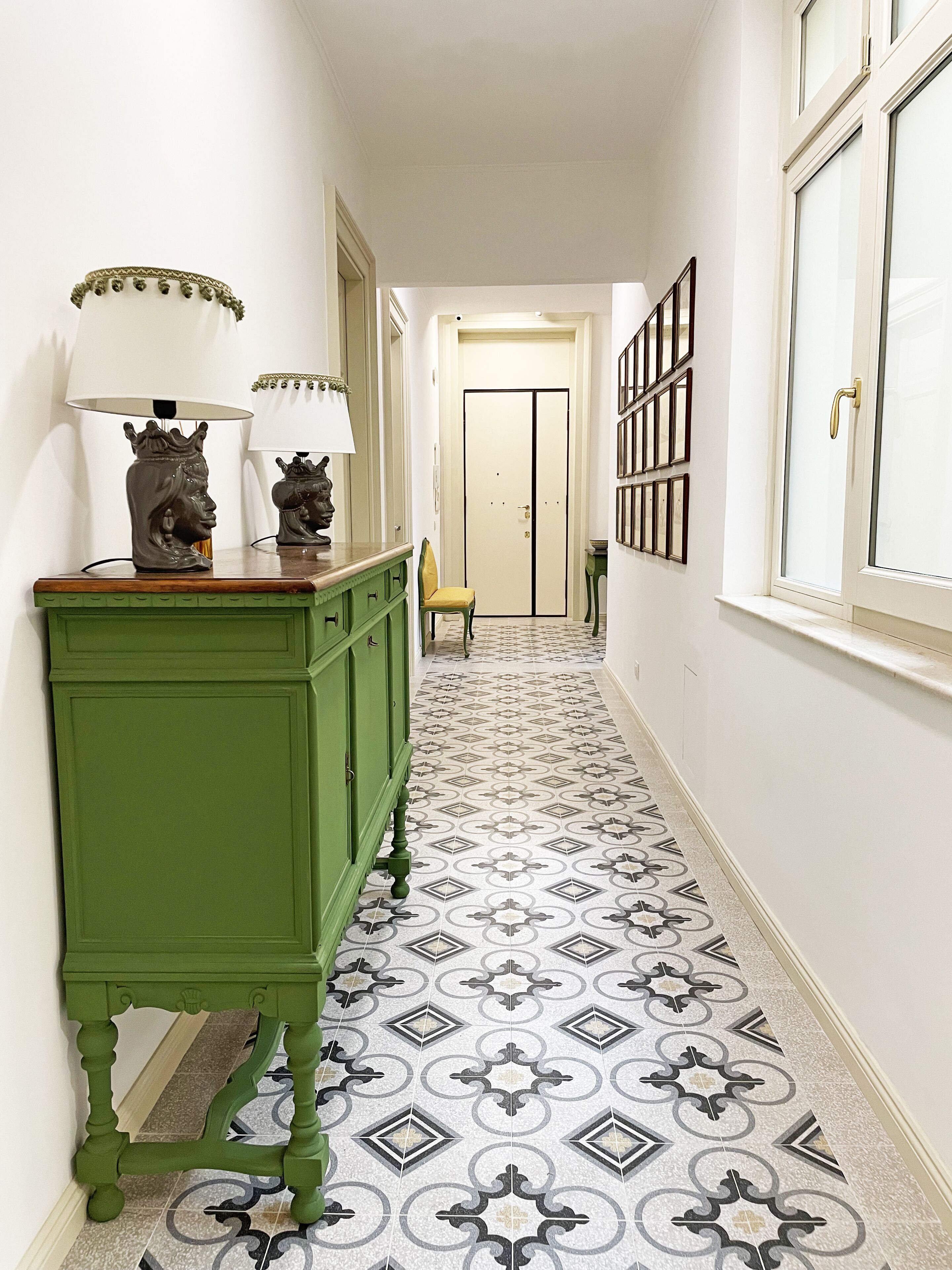 A hallway with patterned floor tiles, a green sideboard with two lamps, and a door at the end.