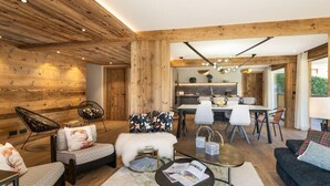 Ein modernes Wohnzimmer mit Holzbalkendecke, einem Essbereich mit weißem Tisch und Stühlen sowie einer Küche mit dunkler Arbeitsplatte.