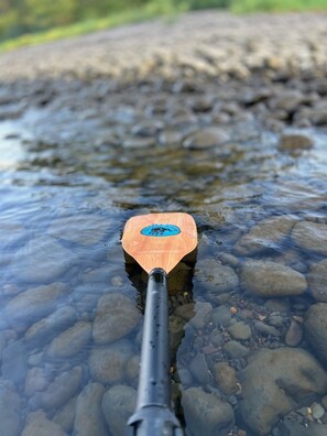 A close-up of a wooden paddle with a logo, floating on clear water with visible pebbles.