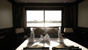 A room with a large window overlooking a body of water, a sofa with cushions, a table with a folded cloth, and two lamps.