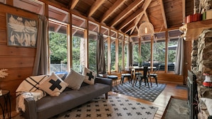 A cozy cabin interior with a wooden ceiling, large windows, a sofa with patterned cushions, a dining area, and a stone wall. - Clerestory Cottage (Big Bear Lake)