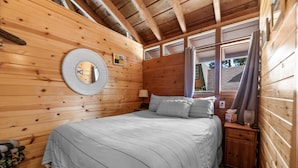 A wooden cabin bedroom with a bed, a nightstand, a lamp, a mirror, and a window with curtains. - Clerestory Cottage (Big Bear Lake)