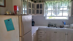 Una cocina con armarios blancos, un refrigerador, un ventanal con cortinas translúcidas y un lavamanos.