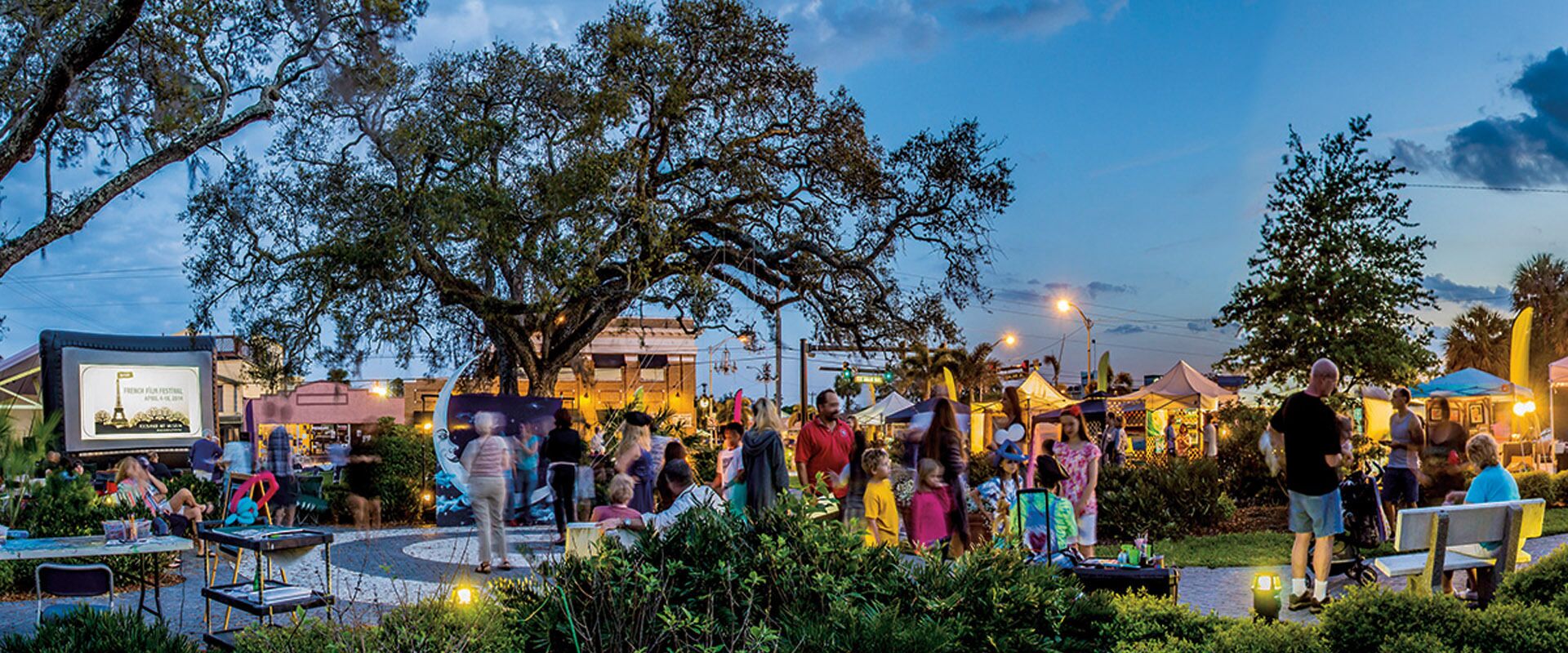 An outdoor event with people gathered around a stage, surrounded by trees and tents.