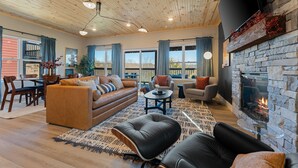 A spacious living room with a stone fireplace, wooden ceiling, and large windows.