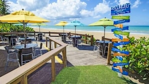 Un ristorante sulla spiaggia con posti a sedere all'aperto, un pontile in legno e un cartello con indicazioni.