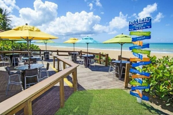 A beachside restaurant with outdoor seating, a wooden deck, and a signpost with destination names. - Beachfront Boutique Villa @ Wyndham Rio Mar Resort (Mameyes II)