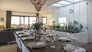 A dining area with a long table set for a meal, a chandelier, and a view of the outdoors through large windows.
