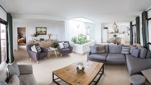 A spacious living area with a gray sofa, a wooden coffee table, and a dining table with chairs. There is a kitchen in the background with a refrigerator and a dining area.