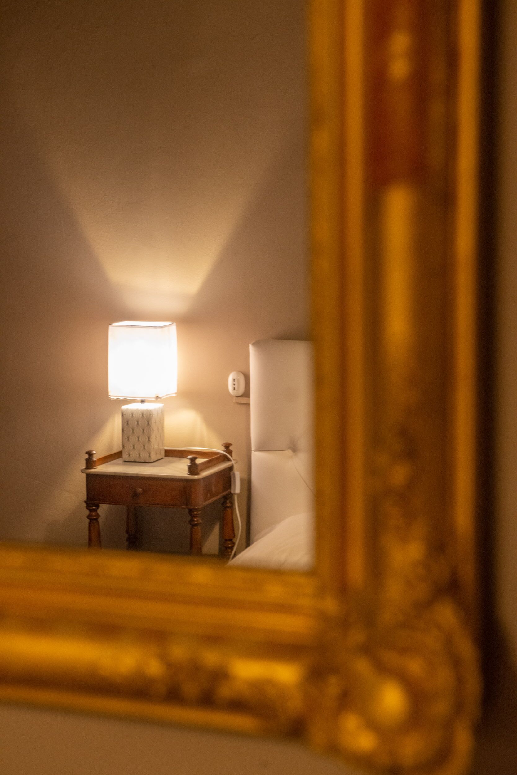 A mirror reflecting a bedside table with a lamp, a wall socket, and a bed.
