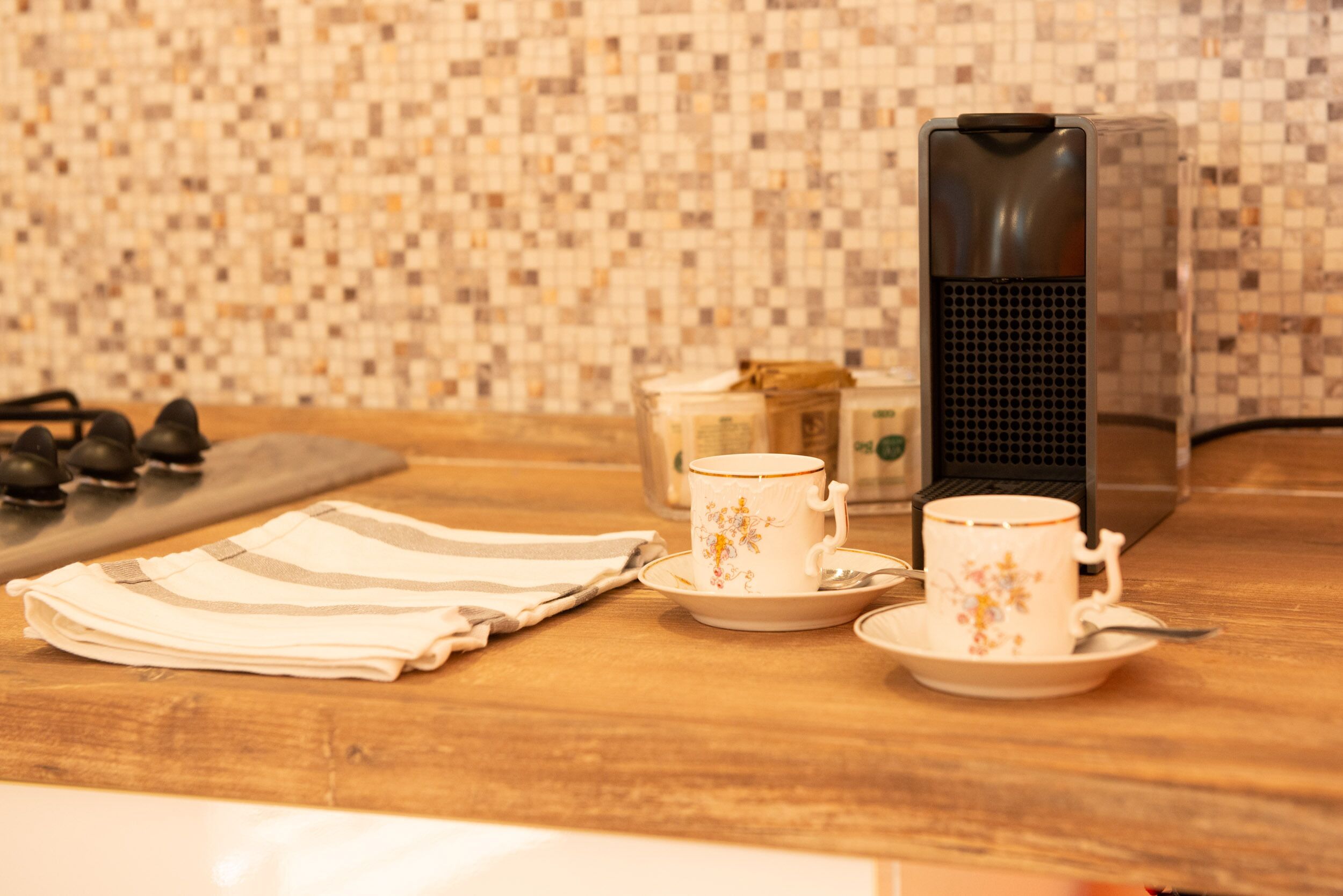 Una encimera de cocina con una cafetera, dos tazas, una tetera y un paño a rayas.