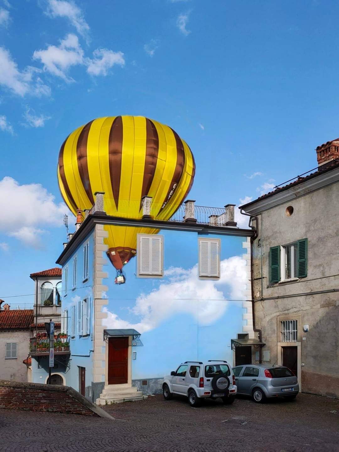 Een gebouw met een grote gele en bruine luchtballon op het dak, geparkeerde auto's ervoor, en een helderblauwe lucht met wolken.
