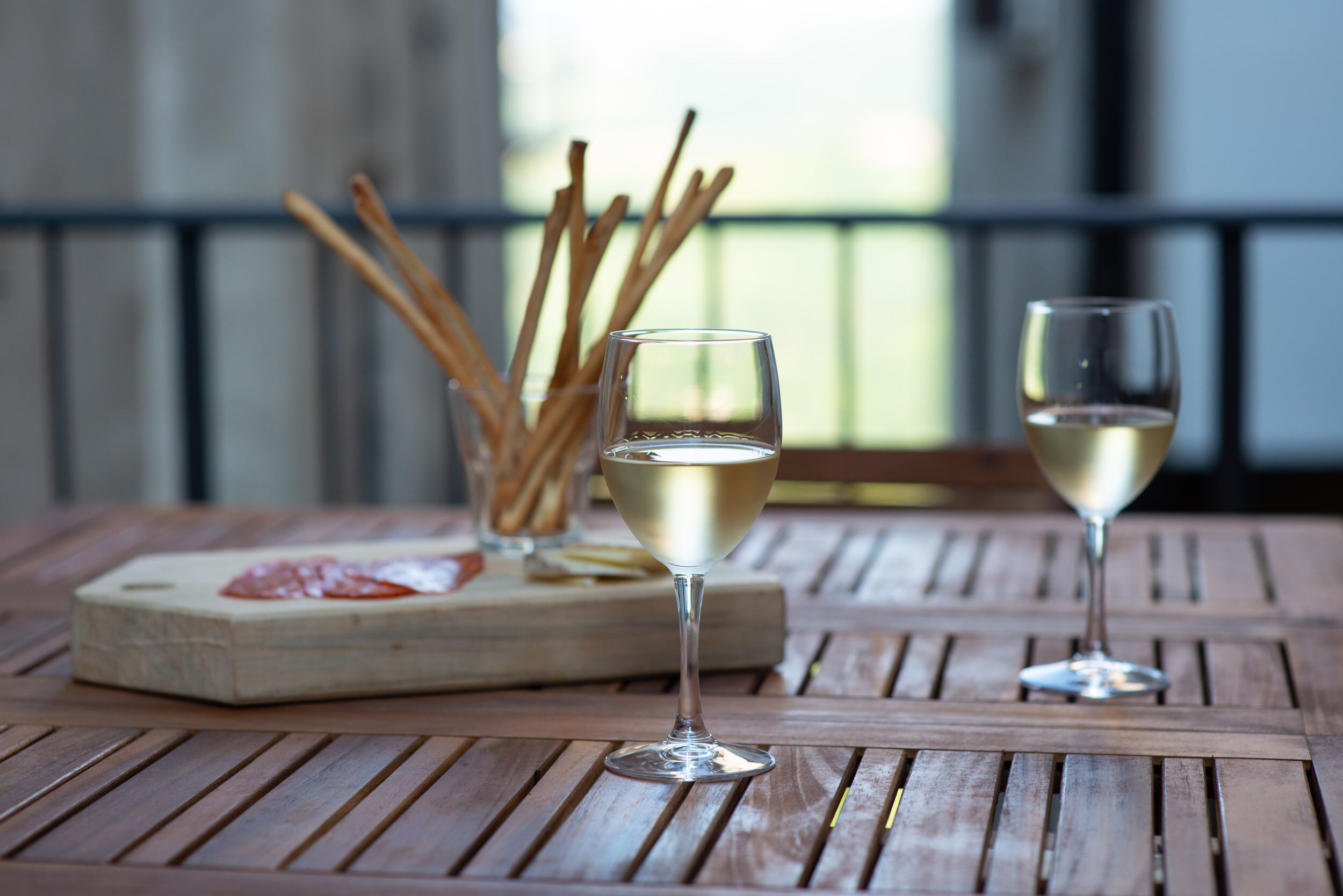 A wooden table with two glasses of white wine and a wooden board with food and sticks.
