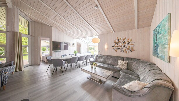 A spacious living area with a grey sofa, dining table, and chairs, wooden flooring, and a large window. - Ferienhaus Ostseele mit Whirlpool, Dampf- und Normale Sauna Sowie Kamin (Brodersby)