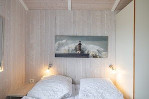 A room with wooden panel walls, a bed with white bedding, two wall-mounted lamps, and a painting of a lighthouse.
