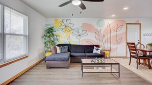 Una sala de estar moderna con un sofá modular, una mesa de centro, una mesa de comedor con sillas, un ventilador de techo y una pared con un mural floral.