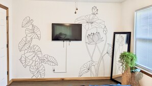 Un dormitorio con un amplio mural floral, un televisor de pantalla plana montado en la pared, una planta en maceta y una cama con cubrecama azul.
